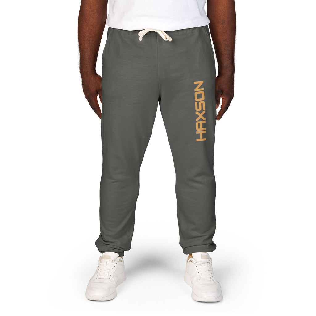 A person wearing dark gray sweatpants with a vertical orange 'HAXSON' logo on the left leg, styled with a white t-shirt and white sneakers against a plain white background.
